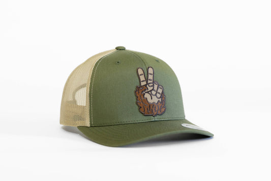 Angled on white, the olive and tan Bigfoot Peace trucker hat shows its furry two finger patch on the front panel.