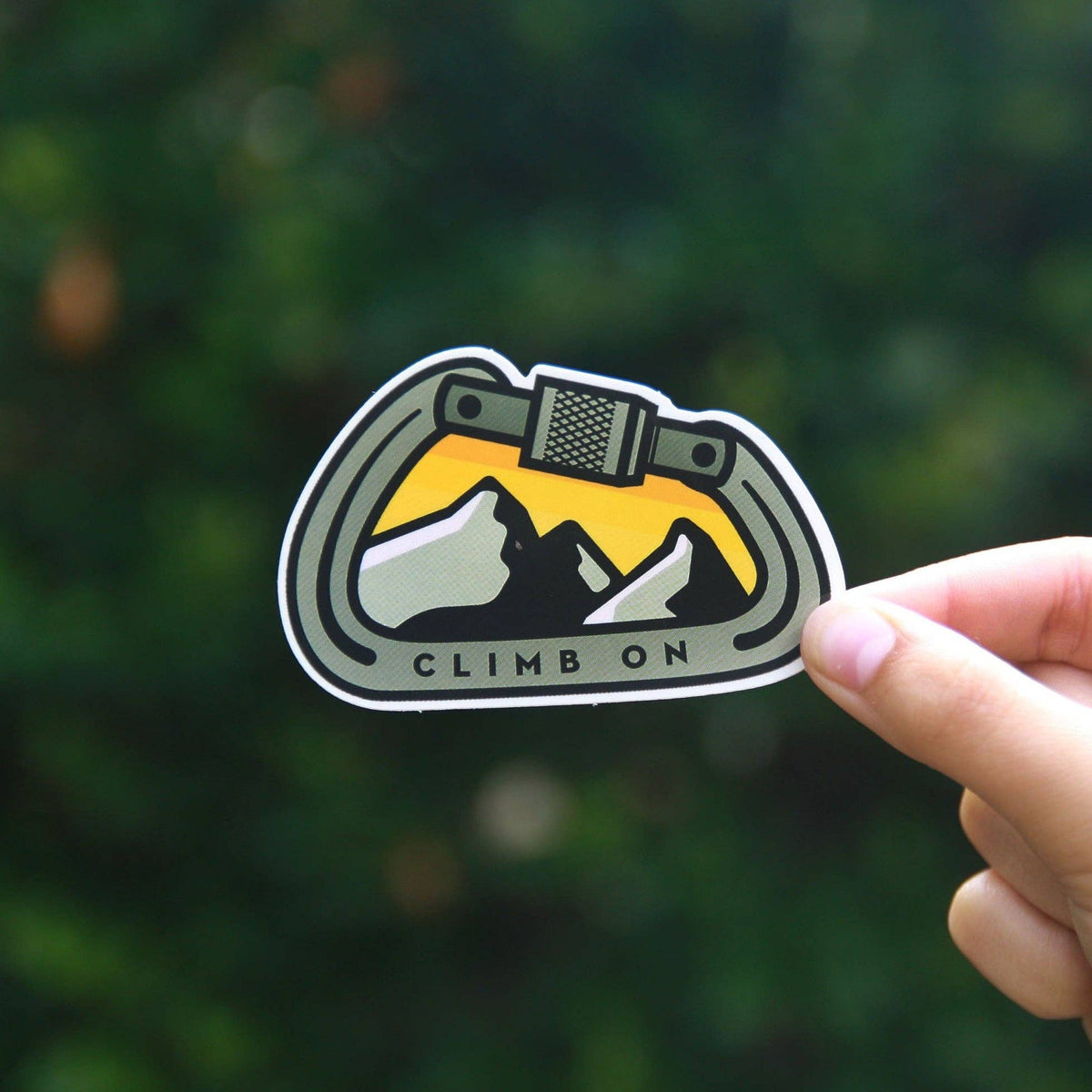 Pinched at one edge, the Climb On Carabiner sticker stands sharp against blurred dark green foliage.