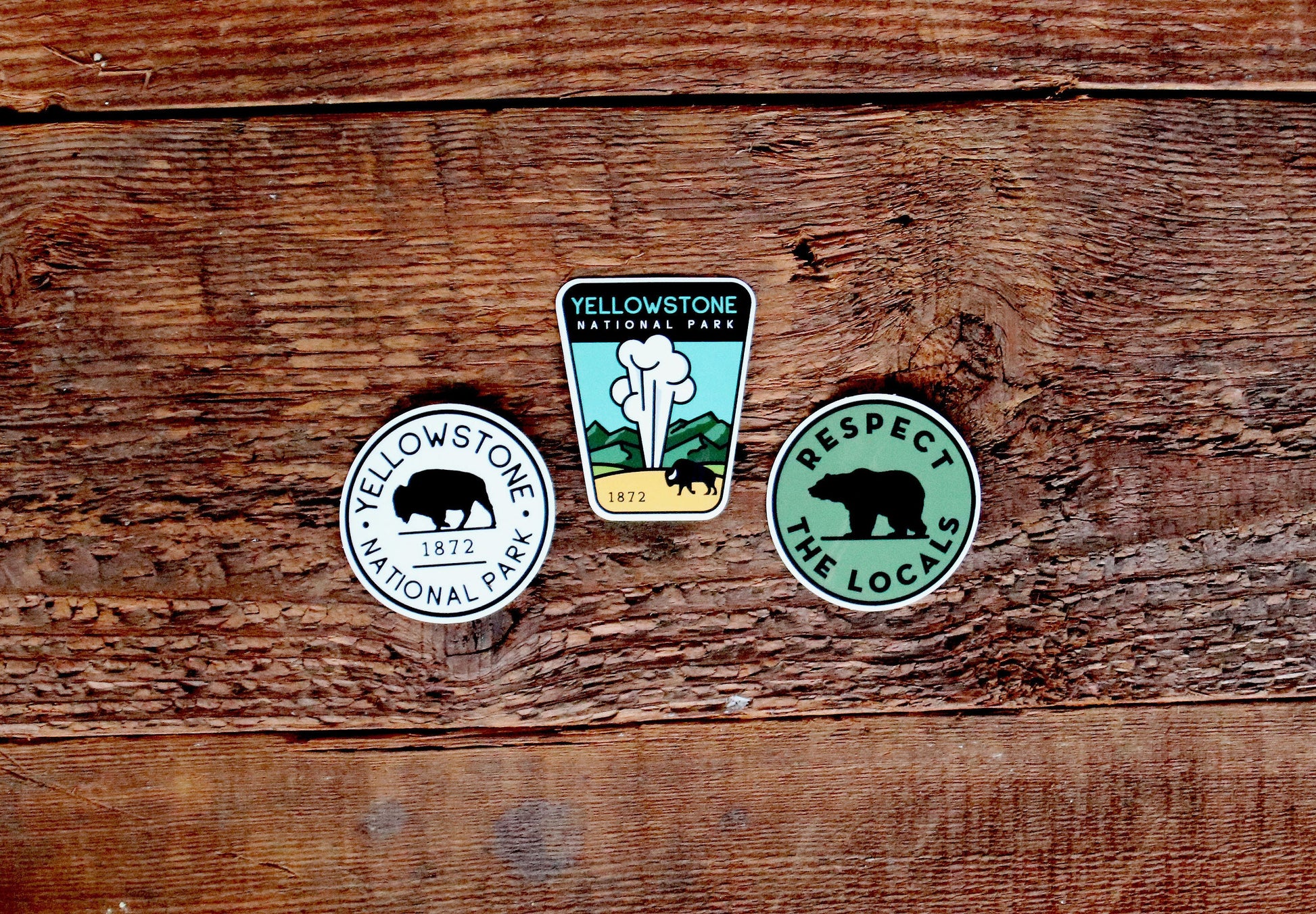Three stickers rest on barnwood, two Yellowstone National Park designs beside a green Respect The Locals bear.