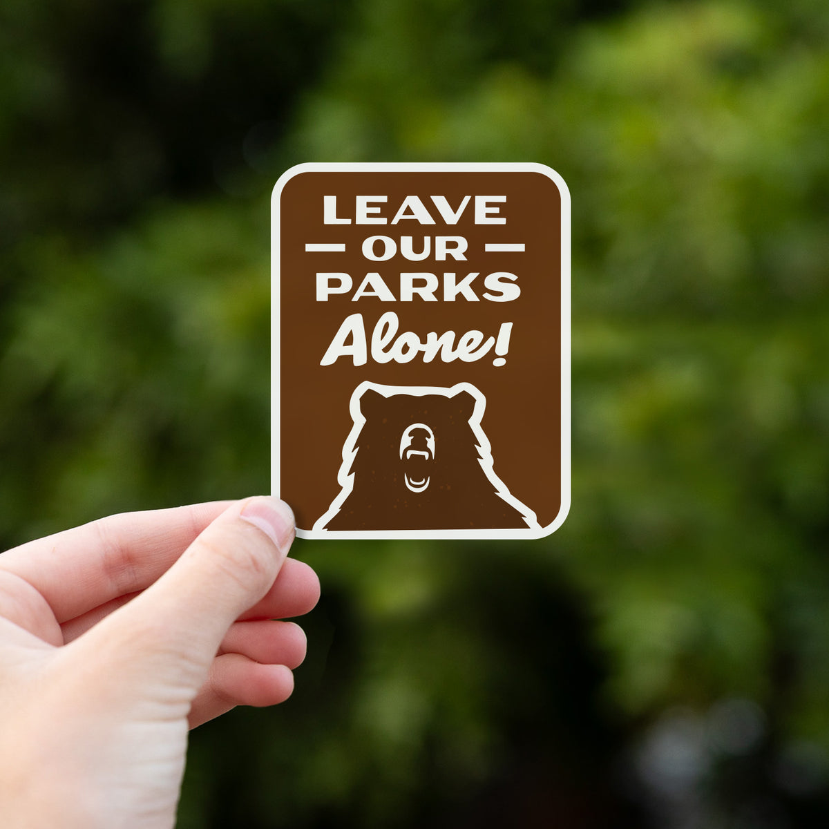 Fingers hold up the Leave Our Parks Alone sticker with soft green foliage filling the background.