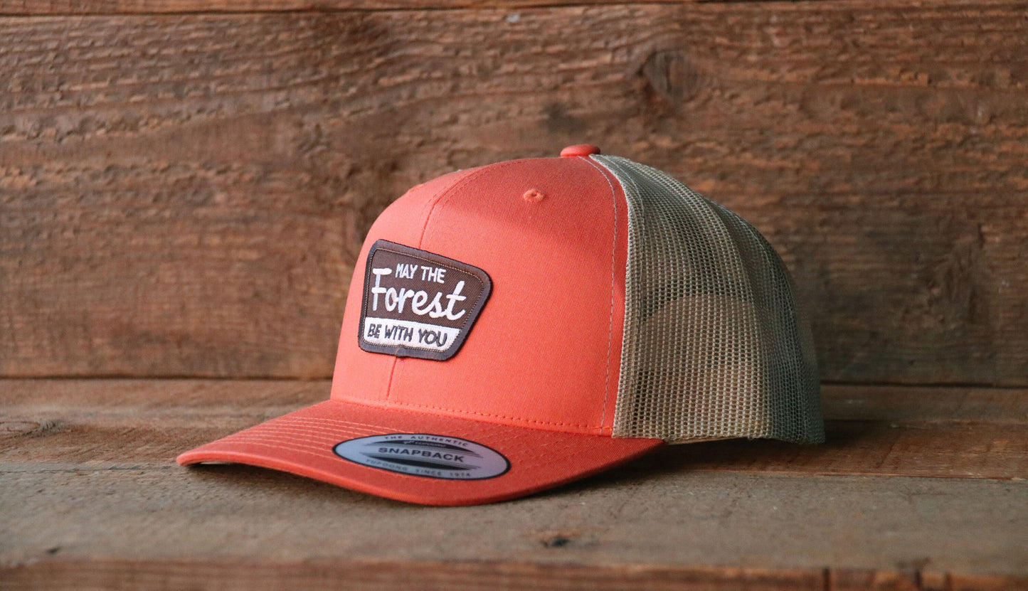 Salmon crown with olive mesh, the May The Forest Be With You trucker hat sits on rough planks by a wood wall.