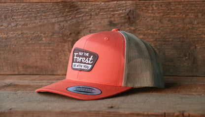 Salmon crown with olive mesh, the May The Forest Be With You trucker hat sits on rough planks by a wood wall.