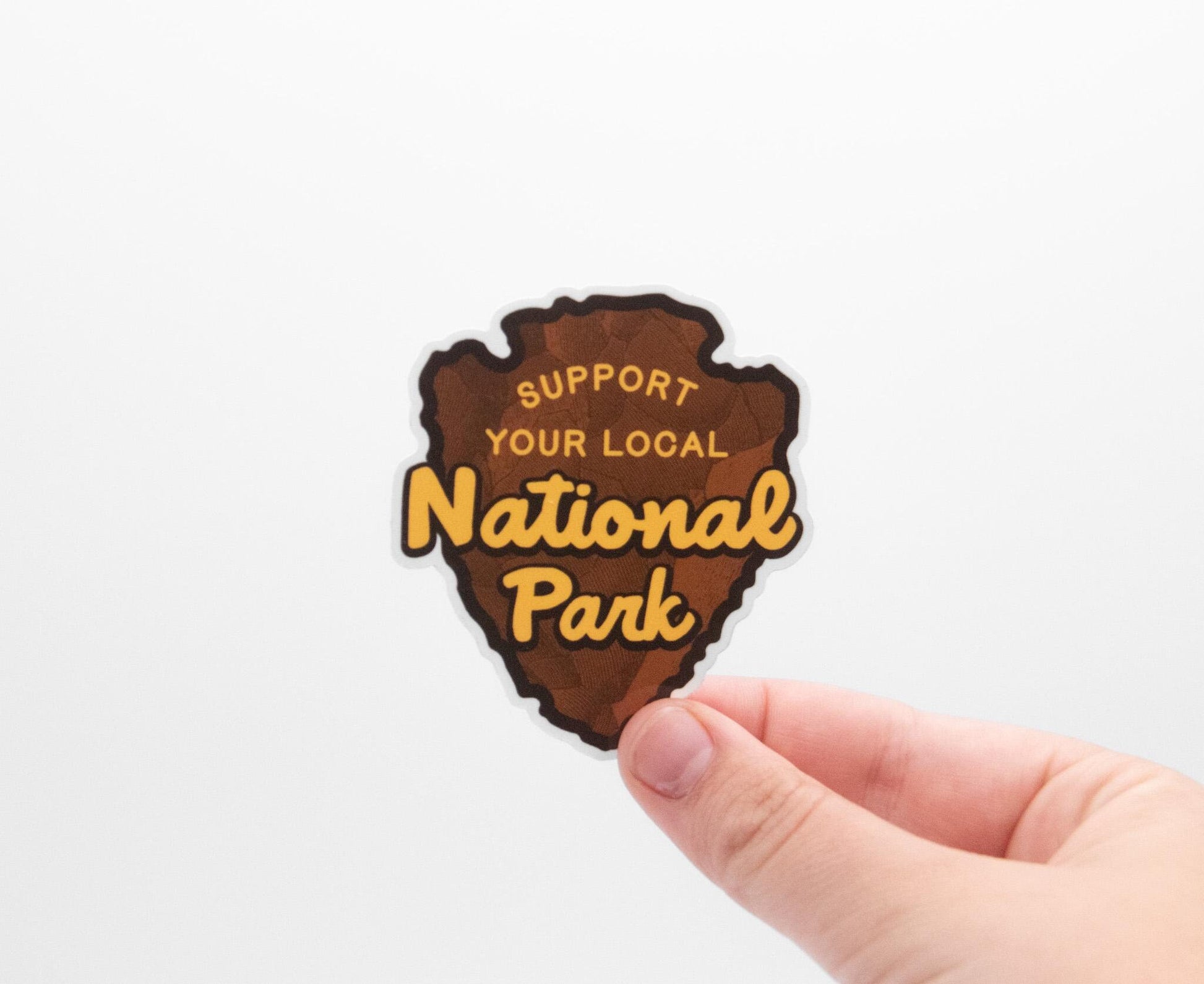 A hand holds the arrowhead shaped Support Your Local National Park sticker, brown with gold lettering, on white.