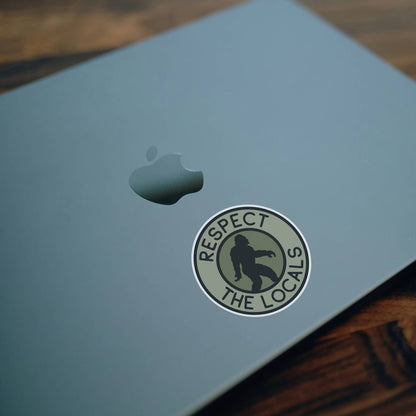 Stuck beside the logo on a pale laptop lid, the Bigfoot Respect The Locals sticker sits over dark wood.