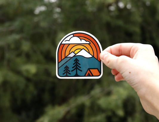 A hand holds the Sunset Campsite sticker against blurred evergreen branches, its orange sun arcing over pines.