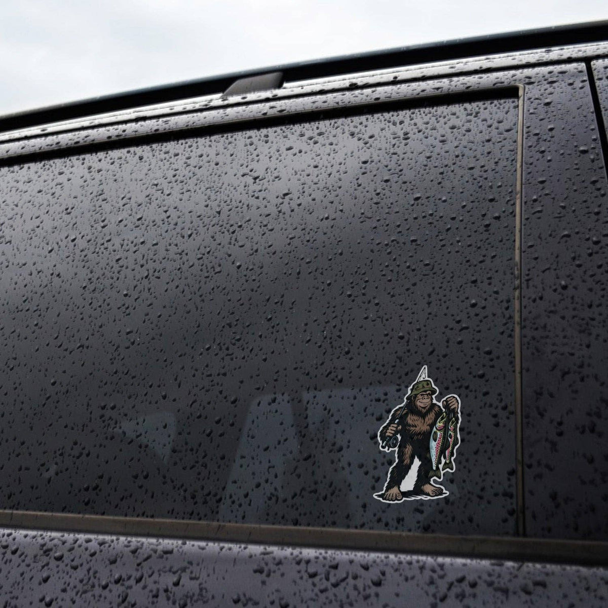 Stuck low on a rain beaded car window, the Bigfoot Fishing sticker sits against dark glass and grey trim.