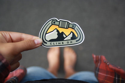 Looking down past bare feet and plaid, a hand holds the Climb On Carabiner sticker over grey asphalt.