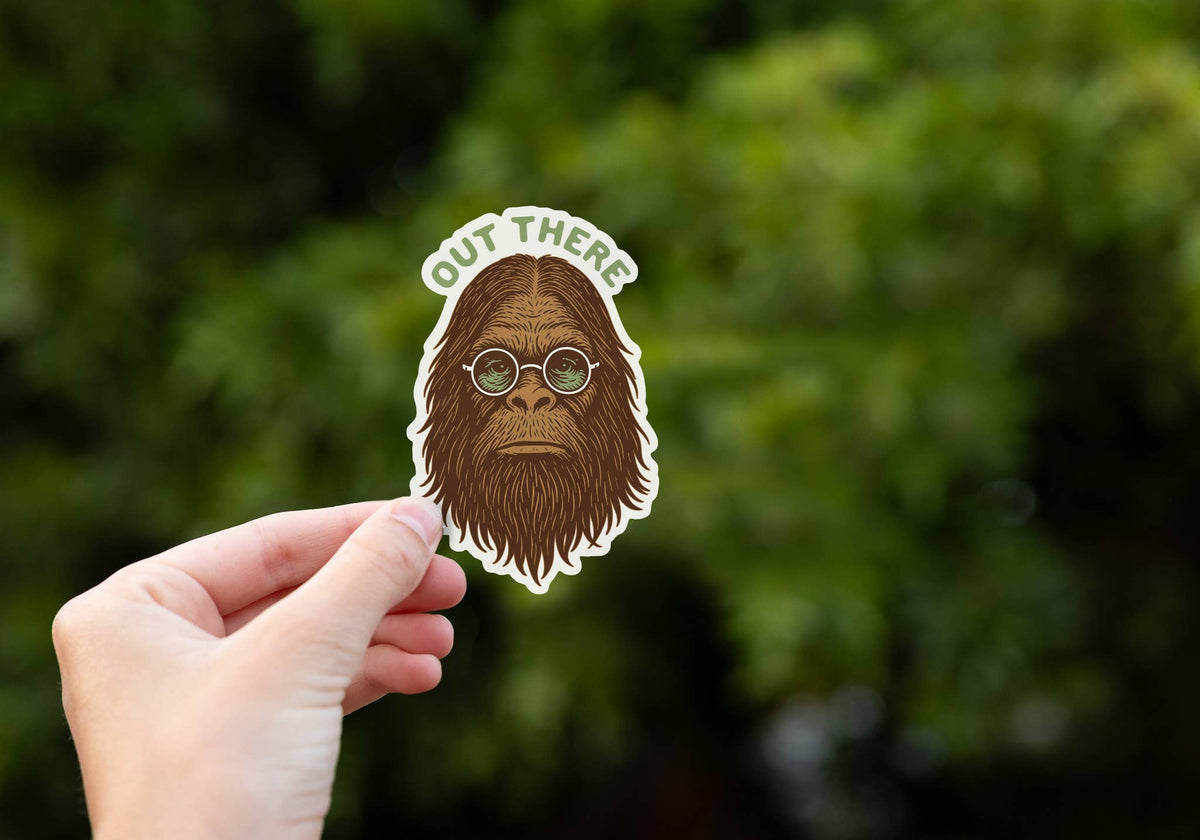 A hand holds the Out There Sasquatch sticker in front of blurred green foliage.