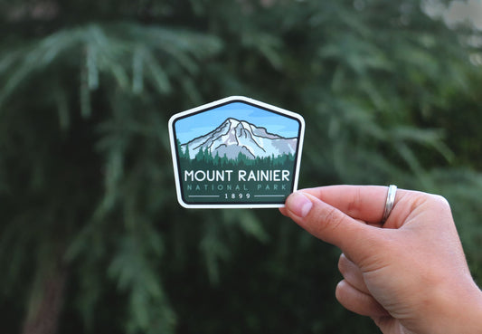 Backed further from the conifer branches, the Mount Rainier National Park sticker shows its snowy peak and 1899 date.