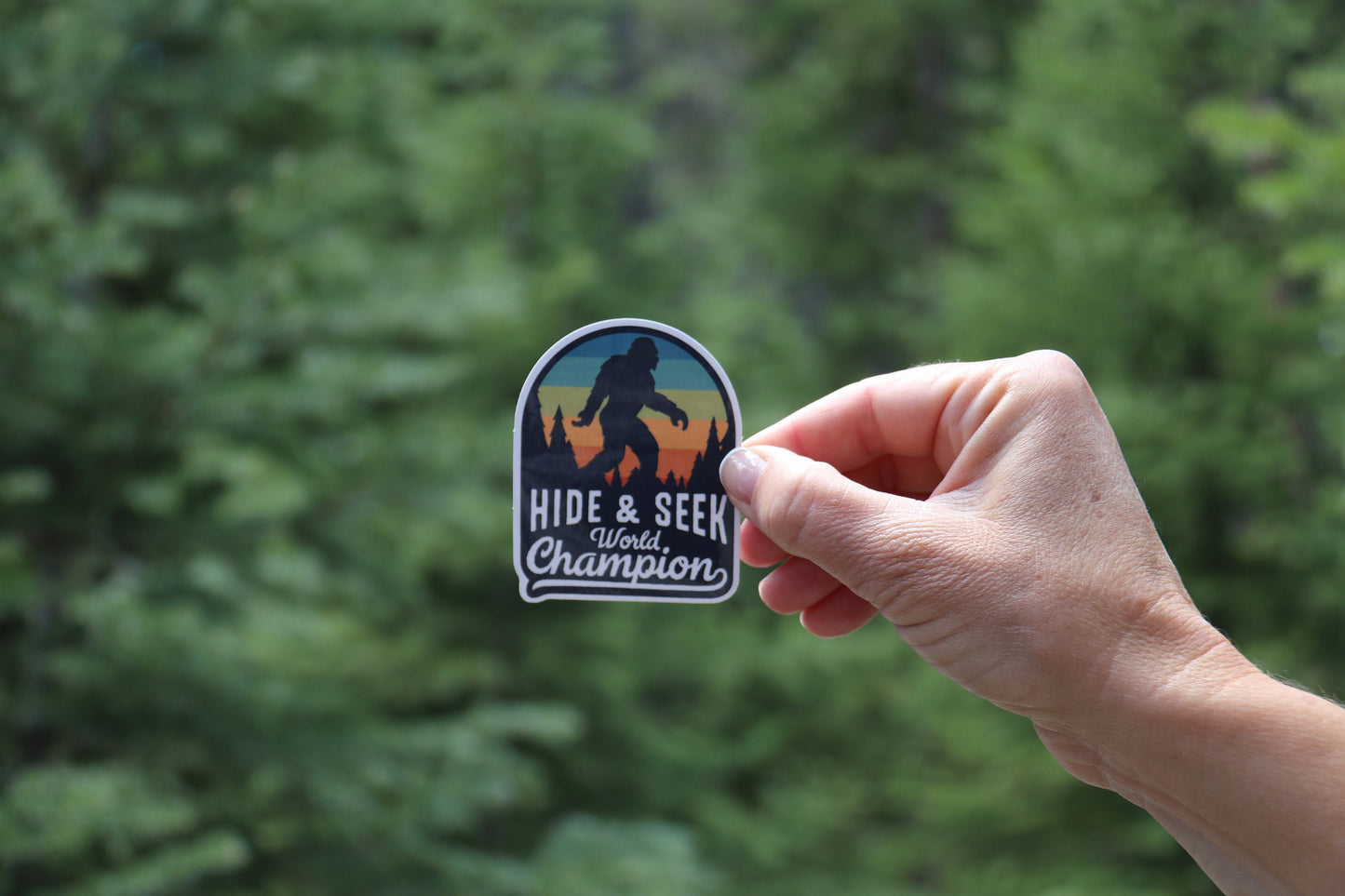 Out among pine trees, a hand holds up the Bigfoot Hide & Seek World Champion sticker by one edge.