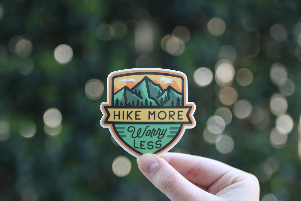 Round spots of light float behind the Hike More Worry Less sticker as a hand holds it near evening foliage.