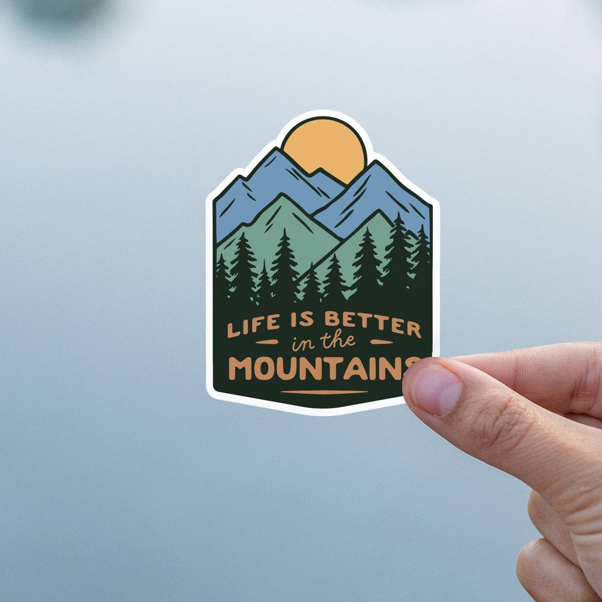 The Life Is Better In The Mountains sticker held up in front of a plain hazy blue backdrop.
