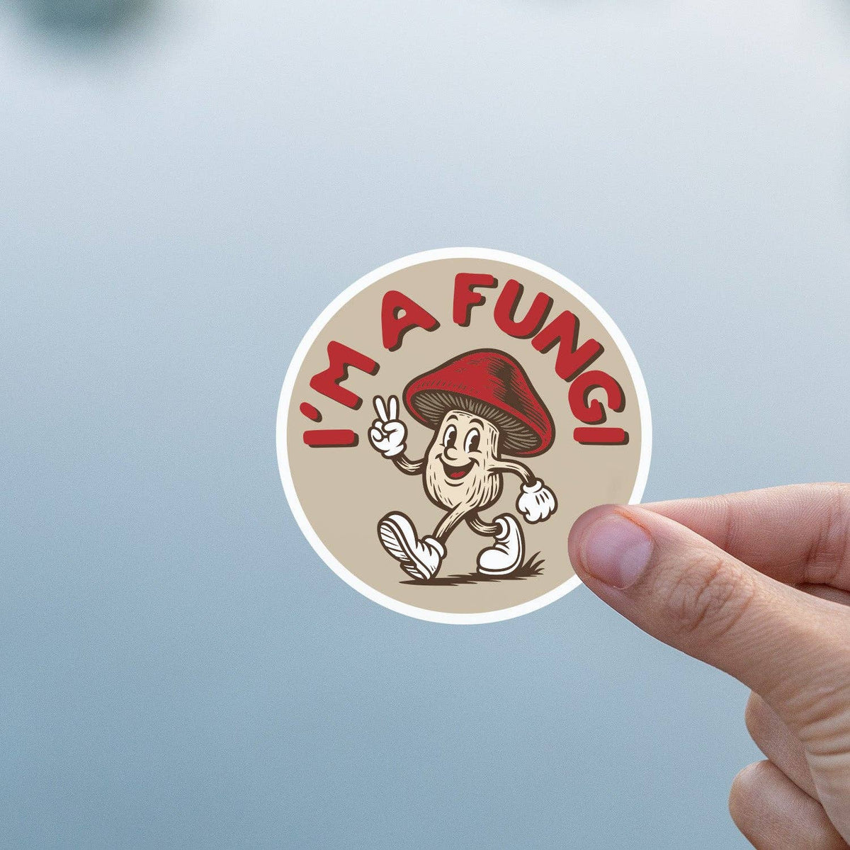 Against a soft blue and grey background, a thumb and finger grip the I'm a Fungi sticker.