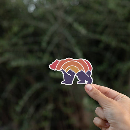 Fingertips lift the Adventure Grizzly Bear sticker in front of a shadowy stand of pine branches.