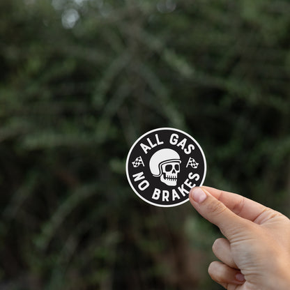 Fingers lift the All Gas No Brakes sticker against a dark blurred hedge of thin branches.