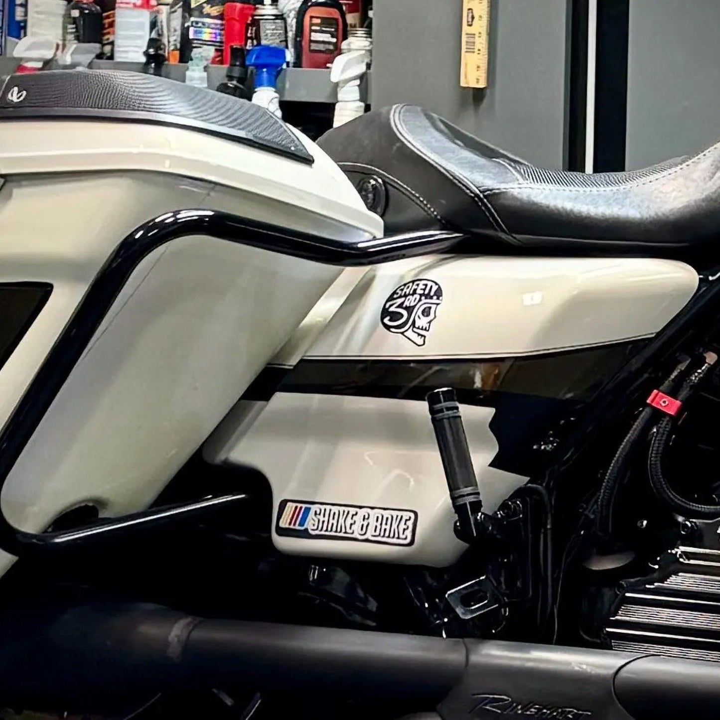 On the cream side panel of a motorcycle in a garage, the Safety 3rd Helmet sticker rides above the engine.