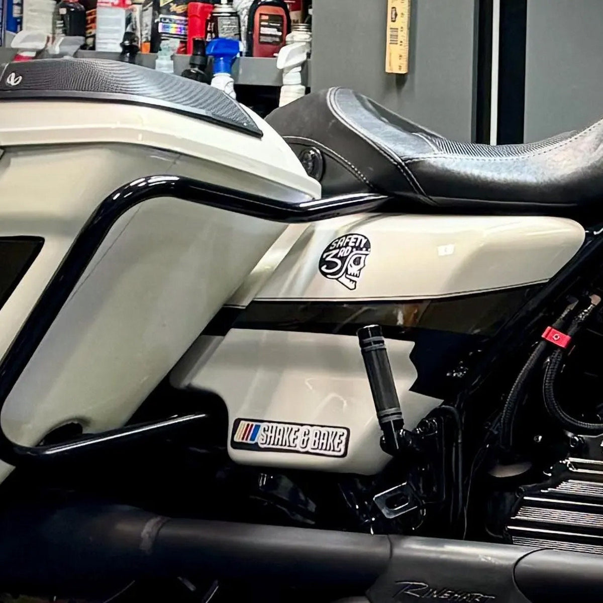 On the cream side panel of a motorcycle in a garage, the Safety 3rd Helmet sticker rides above the engine.