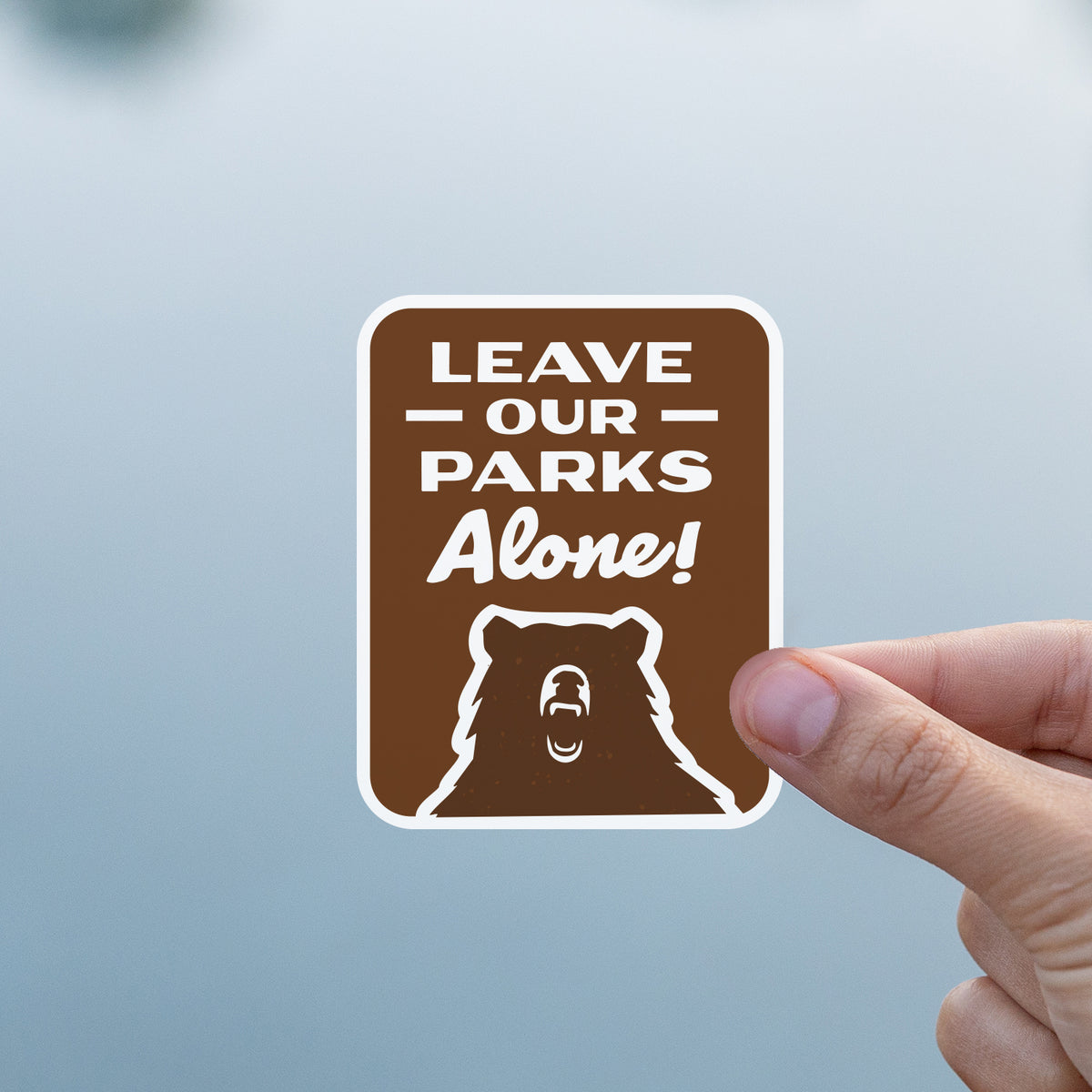A hand presents the Leave Our Parks Alone sticker above the flat grey surface of a lake.