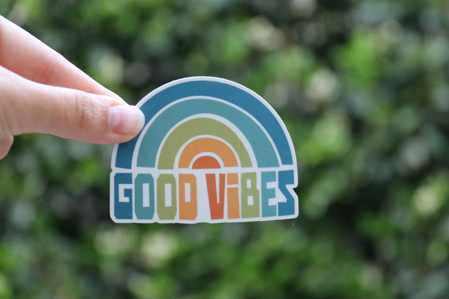One finger balances the Good Vibes sticker at arm's length, small in the frame with a leafy hedge behind.