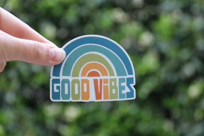 One finger balances the Good Vibes sticker at arm's length, small in the frame with a leafy hedge behind.