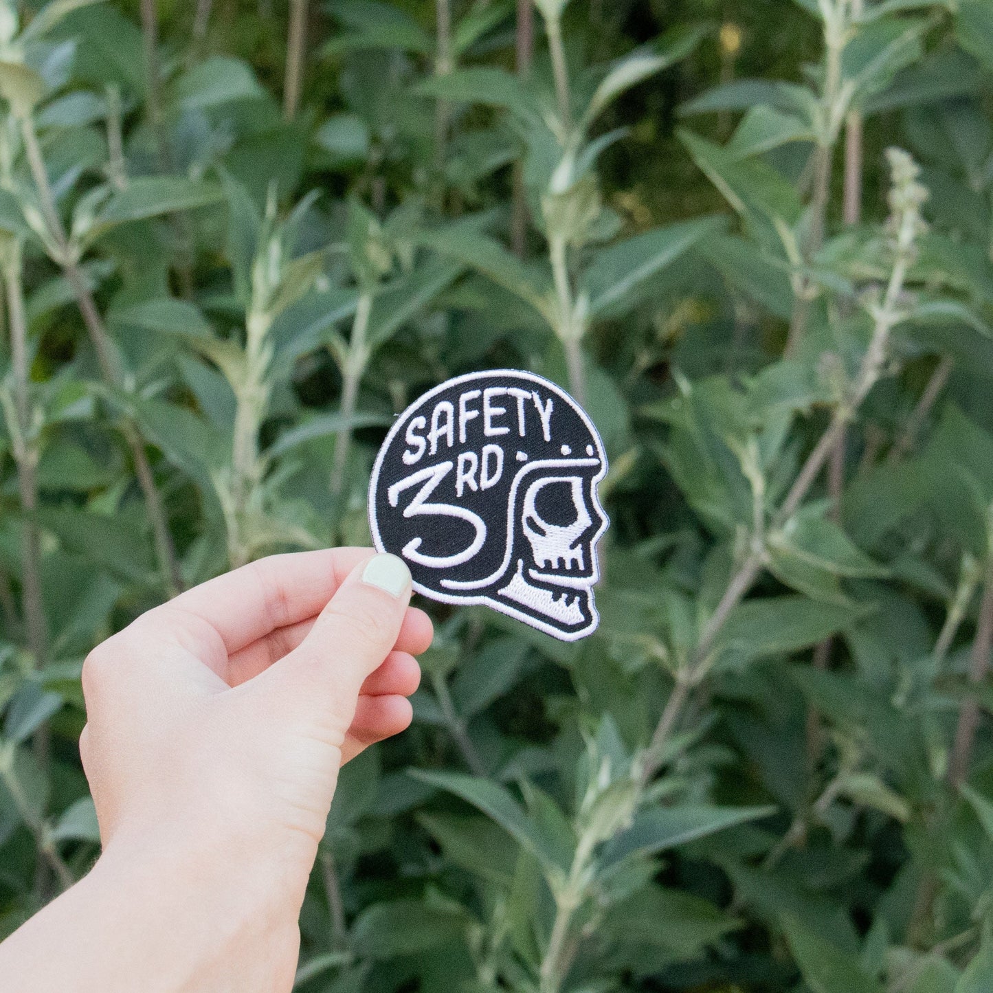 A hand holds the black and white Safety 3rd Helmet Patch, skull in profile, against leafy green branches.