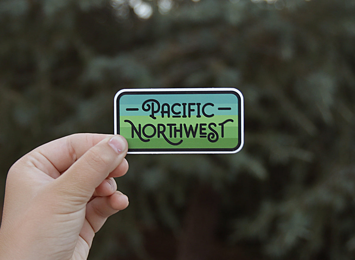 Held further back against blurred conifer branches, the Pacific Northwest sticker reads clearly in black script.