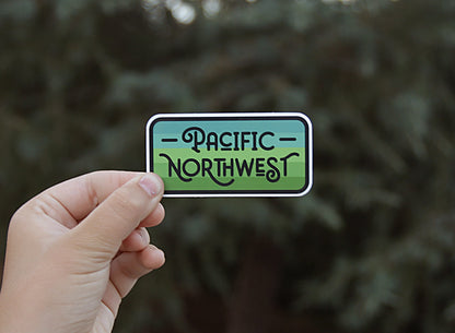 Held further back against blurred conifer branches, the Pacific Northwest sticker reads clearly in black script.