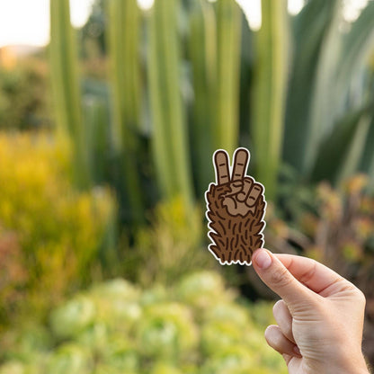 Tall cactus and succulents blur behind a hand holding the Bigfoot Peace sticker at golden hour.