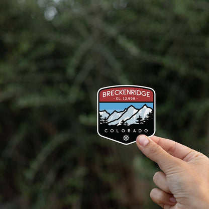 Fingertips lift the Breckenridge Colorado sticker against blurred evergreen branches in shade.