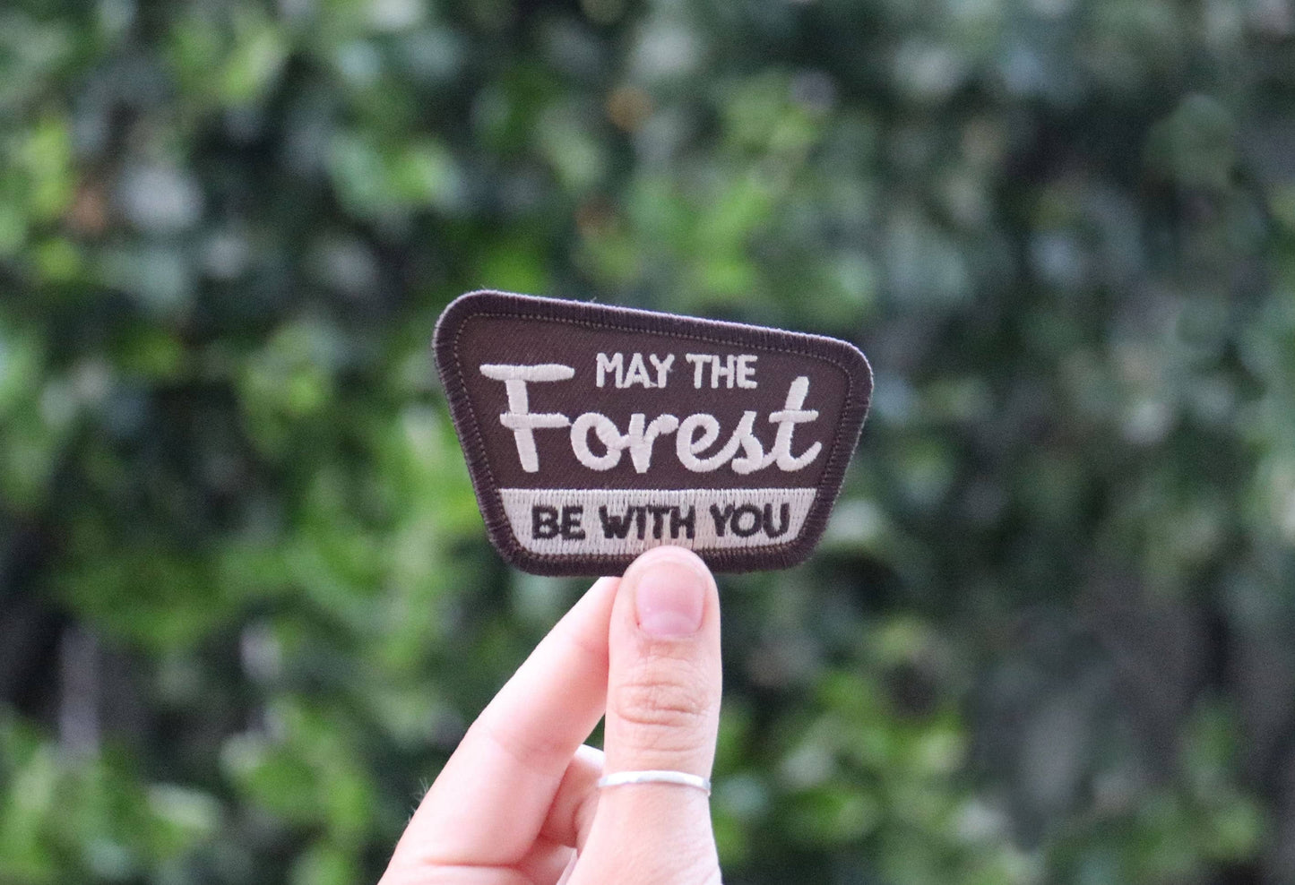 A hand holds the May The Forest Be With You patch up against a blurred wall of green leaves.