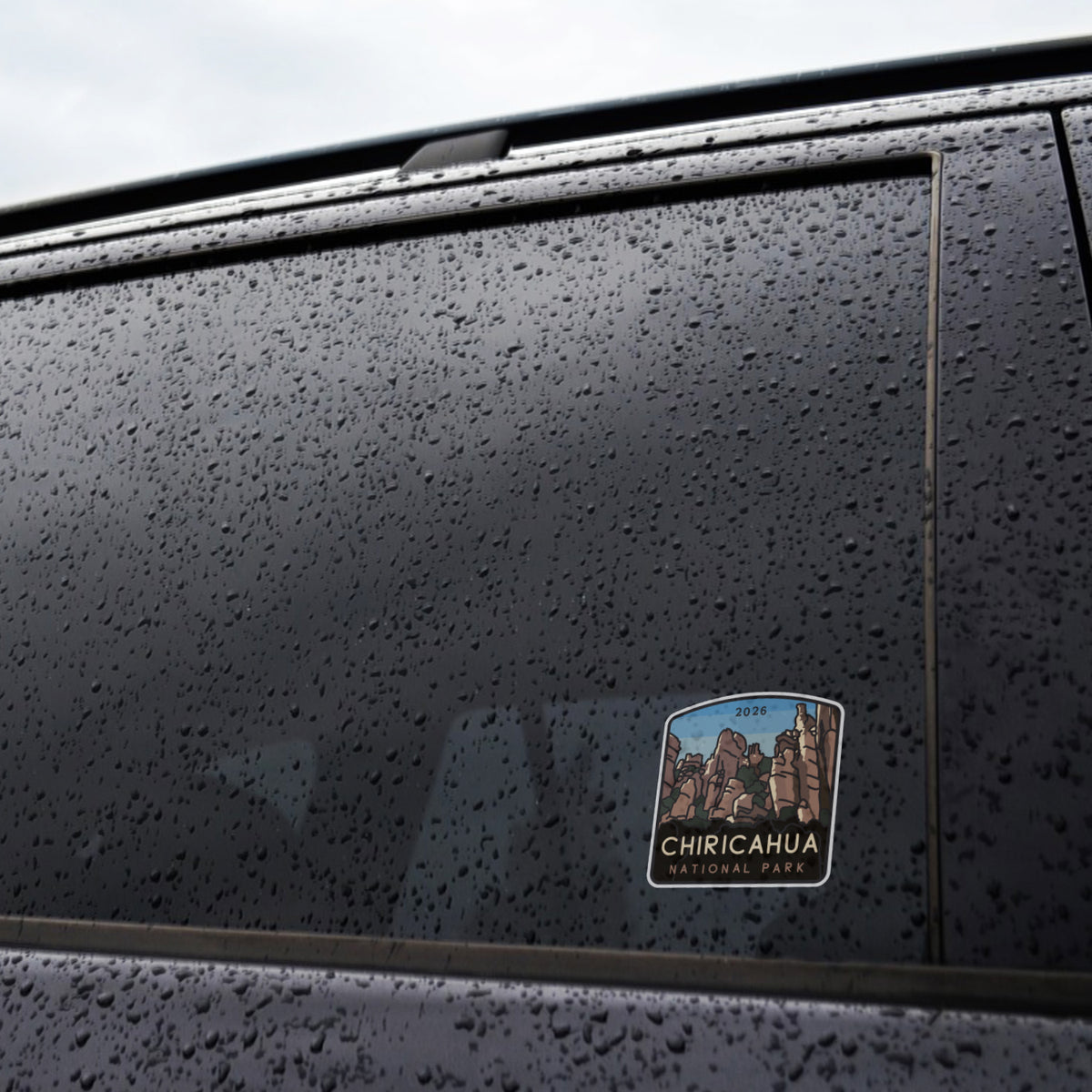 Raindrops cover a dark car window where the Chiricahua National Park sticker clings near the lower corner.
