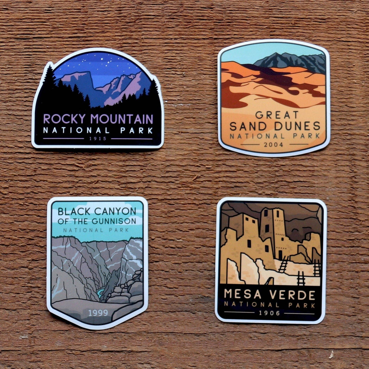 Four stickers from the Colorado National Parks set lie on rough wood, one for each park in the group.