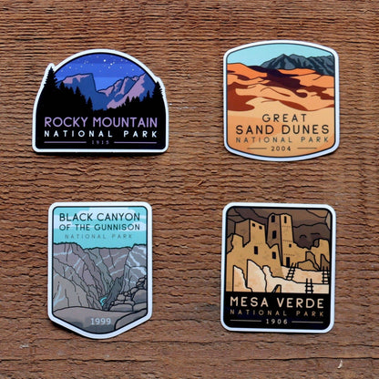 Four stickers from the Colorado National Parks set lie on rough wood, one for each park in the group.