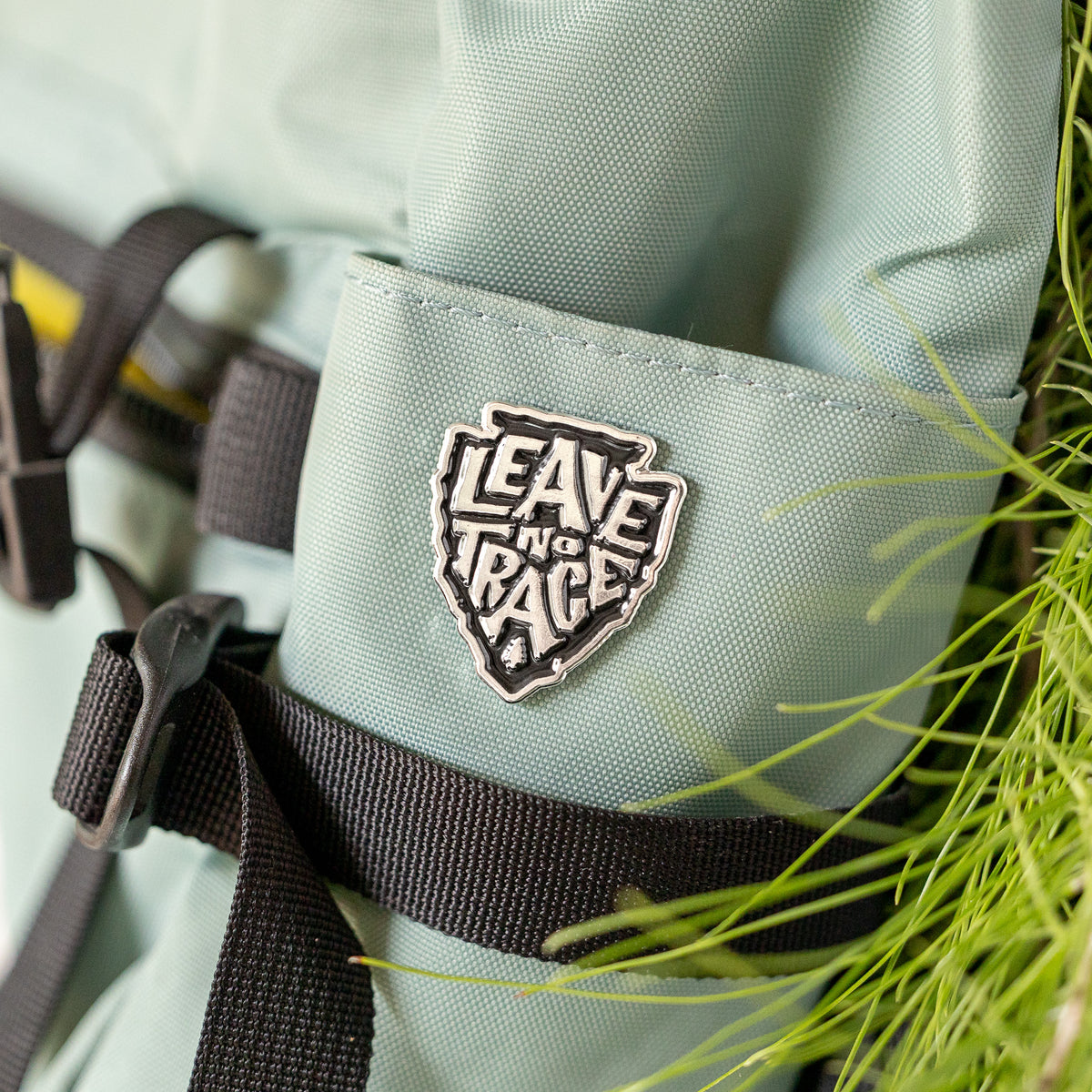 Clipped to the pocket of a pale green backpack, the Leave No Trace pin sits beside tall blades of grass.