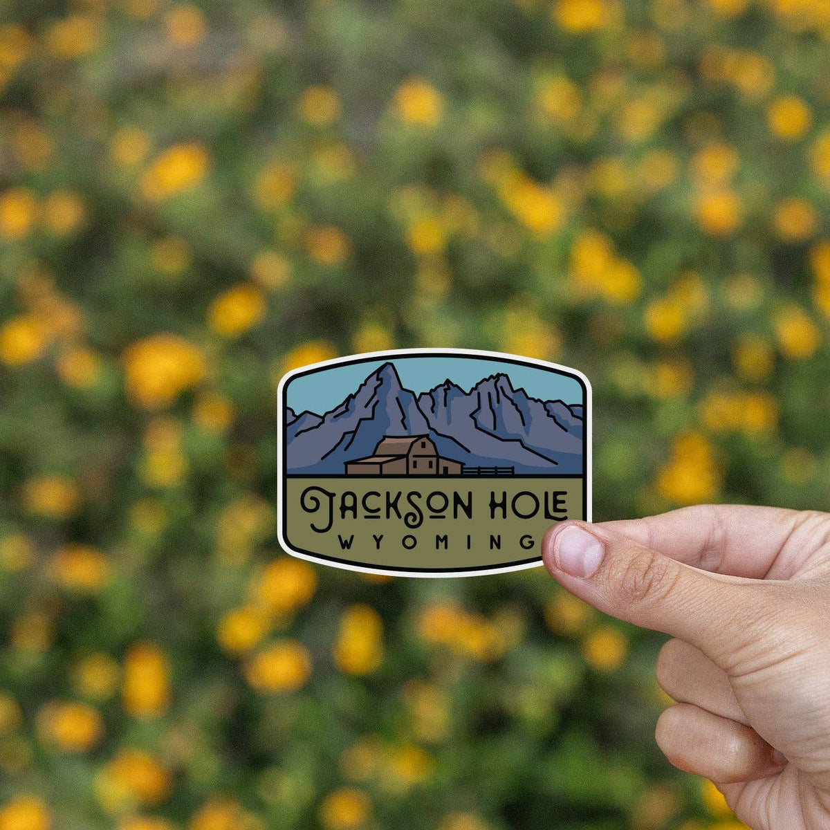 Two fingers hold the Jackson Hole Wyoming sticker in front of a blurred bank of yellow wildflowers.