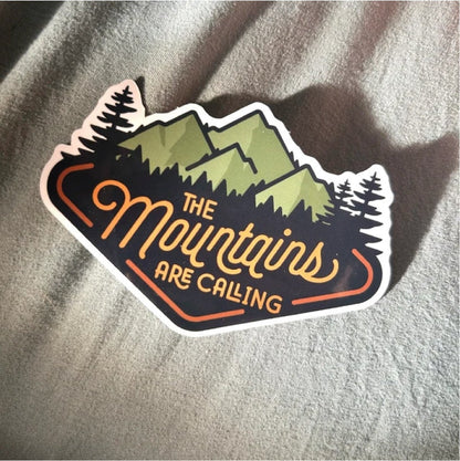 Resting on soft grey fabric in slanted shadow, The Mountains Are Calling sticker catches the light.