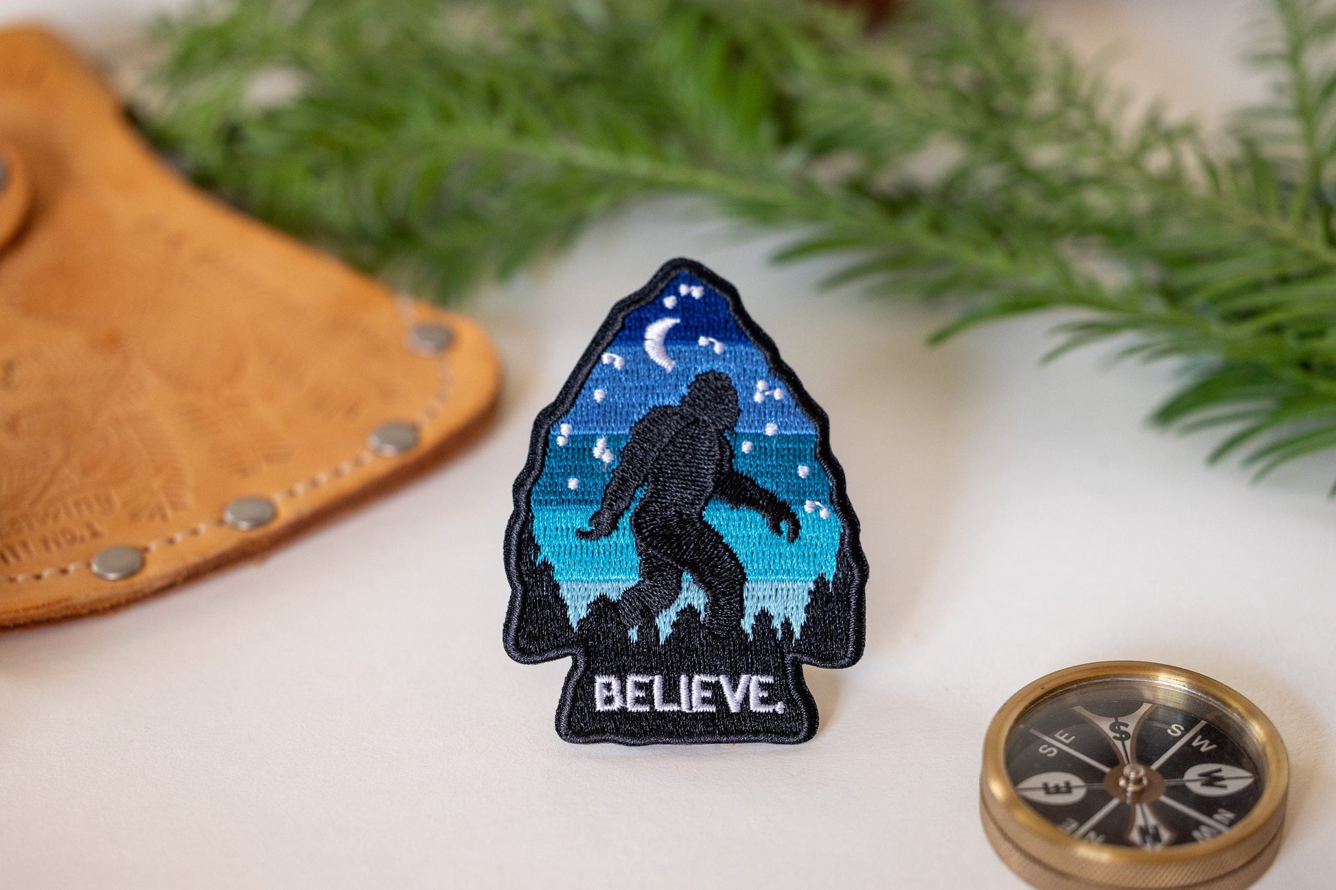 Beside a leather cuff, a brass compass and pine sprigs, the Bigfoot Believe patch stands upright.
