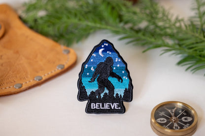 Beside a leather cuff, a brass compass and pine sprigs, the Bigfoot Believe patch stands upright.