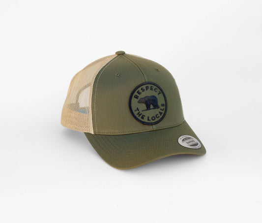 Olive front and tan mesh on the Respect the Locals Bear trucker hat, its round bear patch angled toward the light.