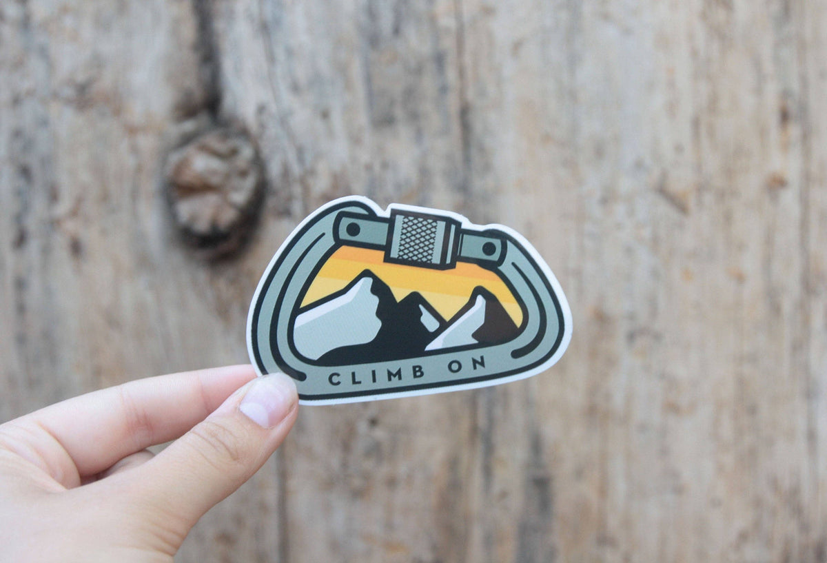 Fingers press the Climb On Carabiner sticker against a weathered wood plank with a dark knot.