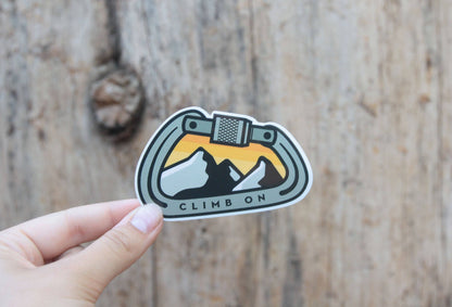 Fingers press the Climb On Carabiner sticker against a weathered wood plank with a dark knot.