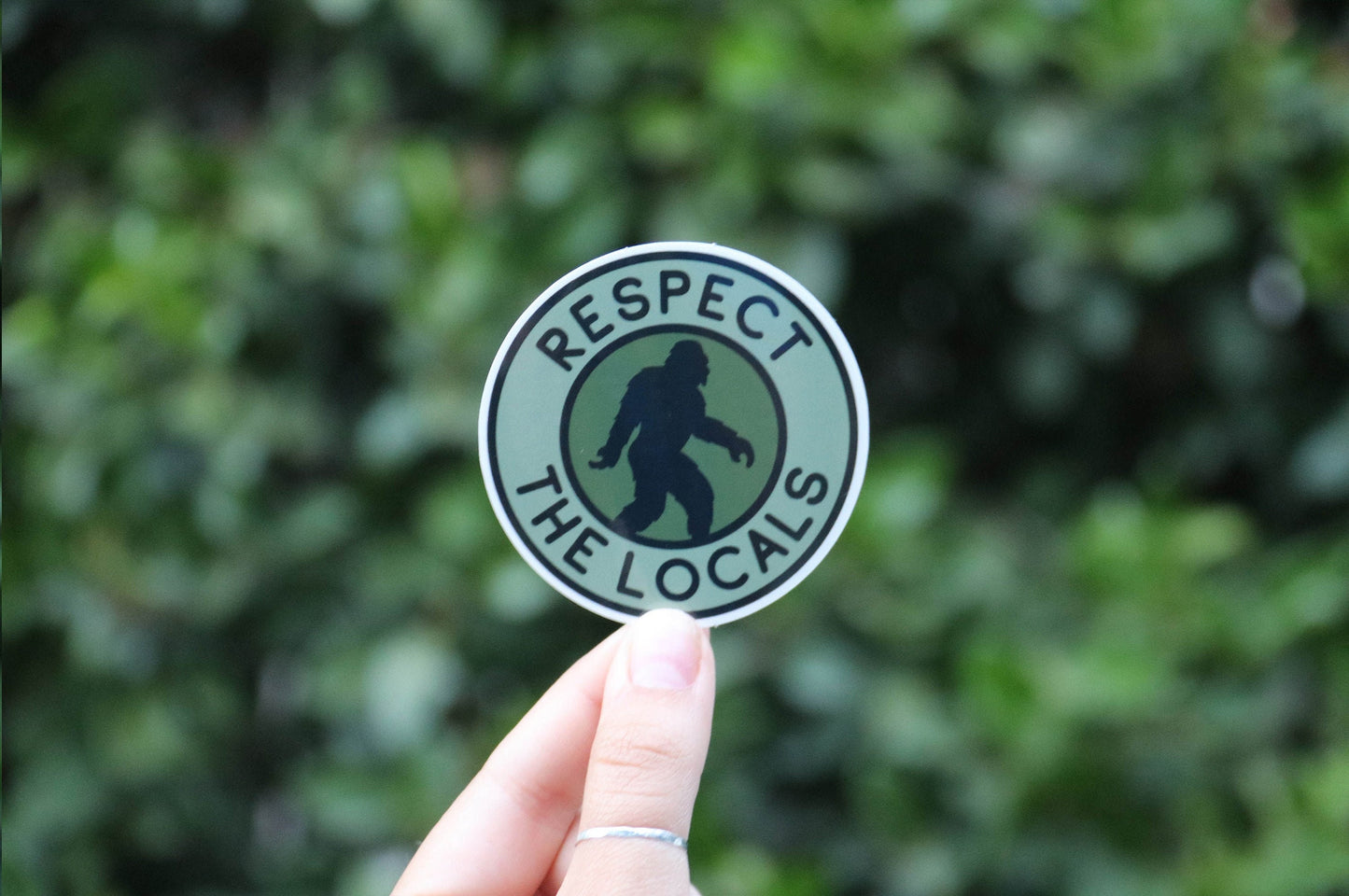 Wider shot of a ringed hand raising the Bigfoot Respect The Locals sticker before a dense green hedge.