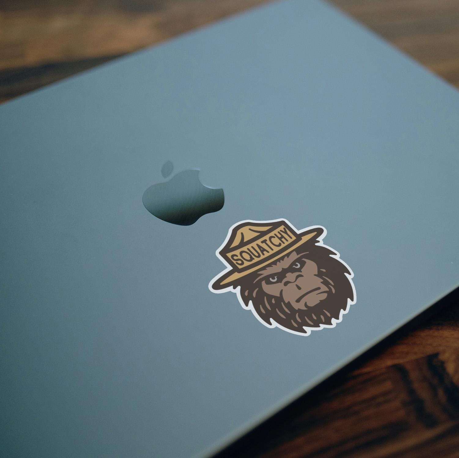 Applied beside the apple on a blue laptop lid, the Squatchy sticker rests over a dark wood desk.