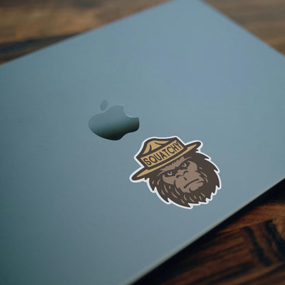 Applied beside the apple on a blue laptop lid, the Squatchy sticker rests over a dark wood desk.
