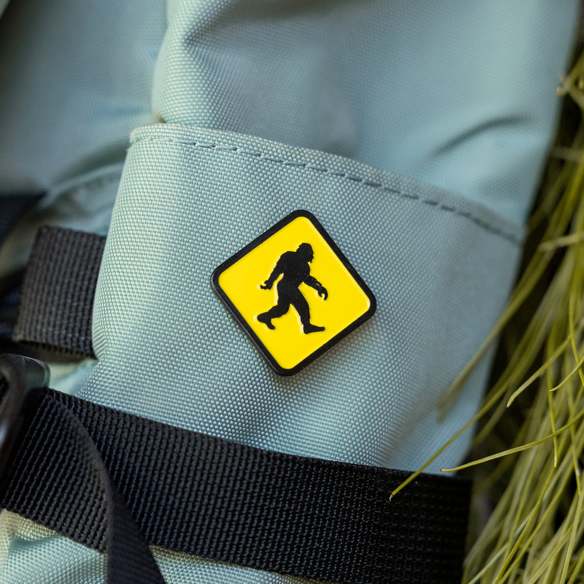 Clipped to the zippered pocket of a grey backpack, the Bigfoot Crossing pin sits beside dried grass stems.