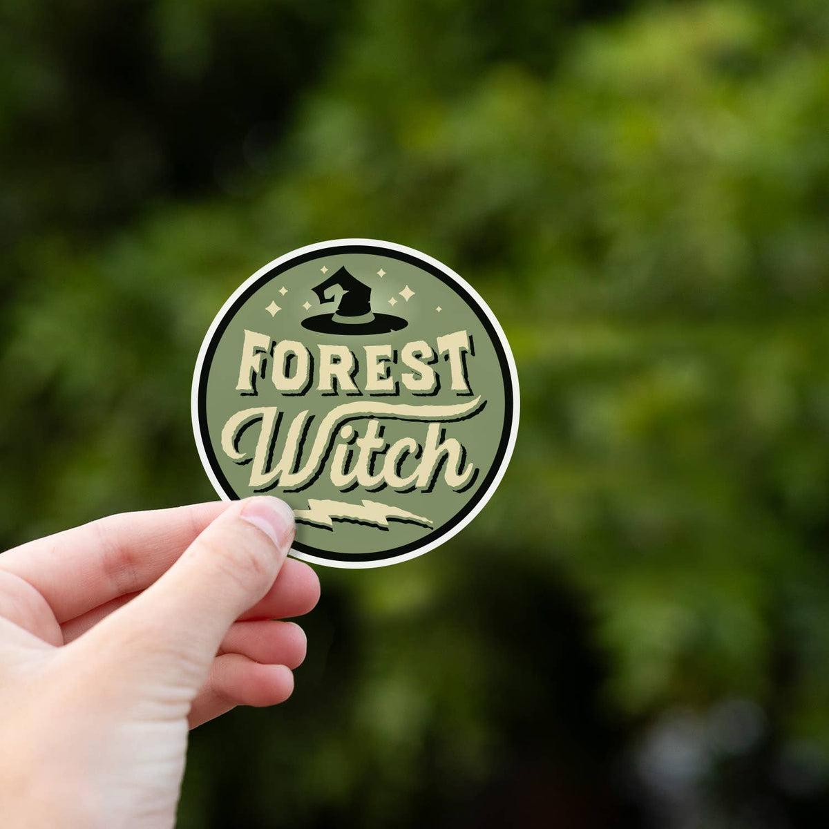 Against dense green leaves, a hand pinches the Forest Witch sticker by its lower edge.
