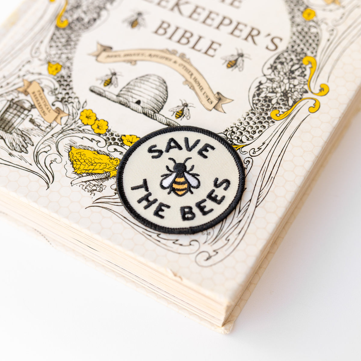 The round Save The Bees patch rests on the corner of The Beekeeper's Bible, its embroidered bee matching the cover art.