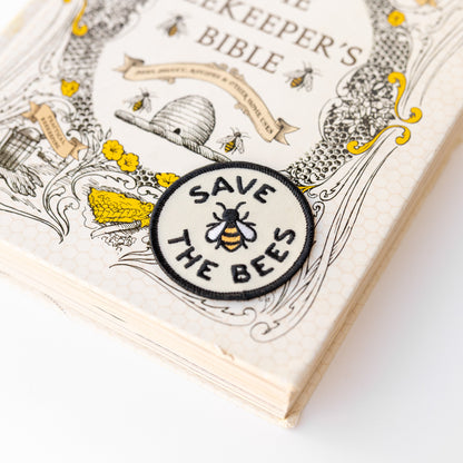 The round Save The Bees patch rests on the corner of The Beekeeper's Bible, its embroidered bee matching the cover art.