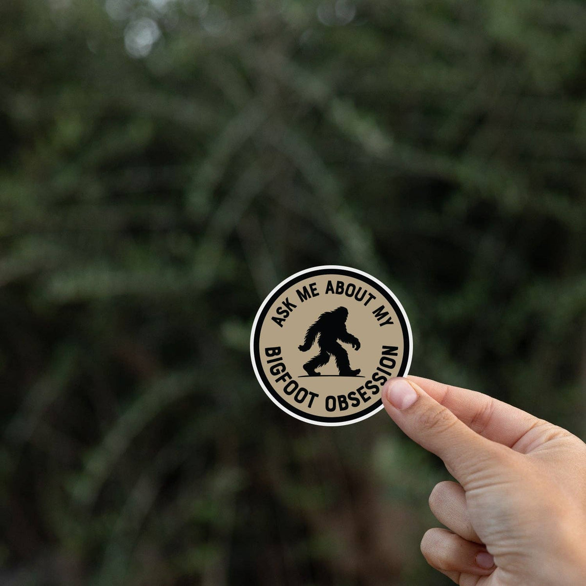 A hand holds the Ask Me About My Bigfoot Obsession sticker in front of blurred evergreen branches.