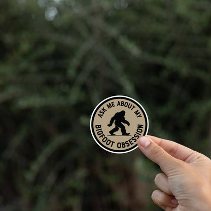 A hand holds the Ask Me About My Bigfoot Obsession sticker in front of blurred evergreen branches.